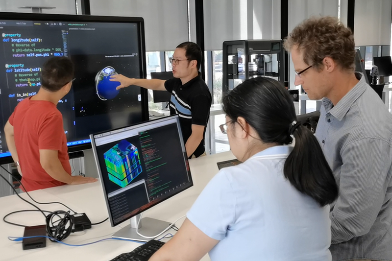 A group of four geospatial scientists consult computer and TV screens, which displays code, colourful cross sections of the earth, and a floating atlas.