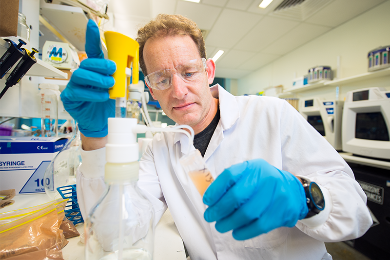 A researcher, wearing a black long sleeved t-shirt covered mostly by a white lab coat, stands in a laboratory. He is surrounded by small miscellaneous machines, scientific vessels, and bags of soil He is holding a small plastic funnel, and appears to be transferring a solution into another vessel, that likely contains some soil within a chemical solution.