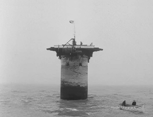 Black and white image of a pillar in the ocean, with a row boat in the foreground