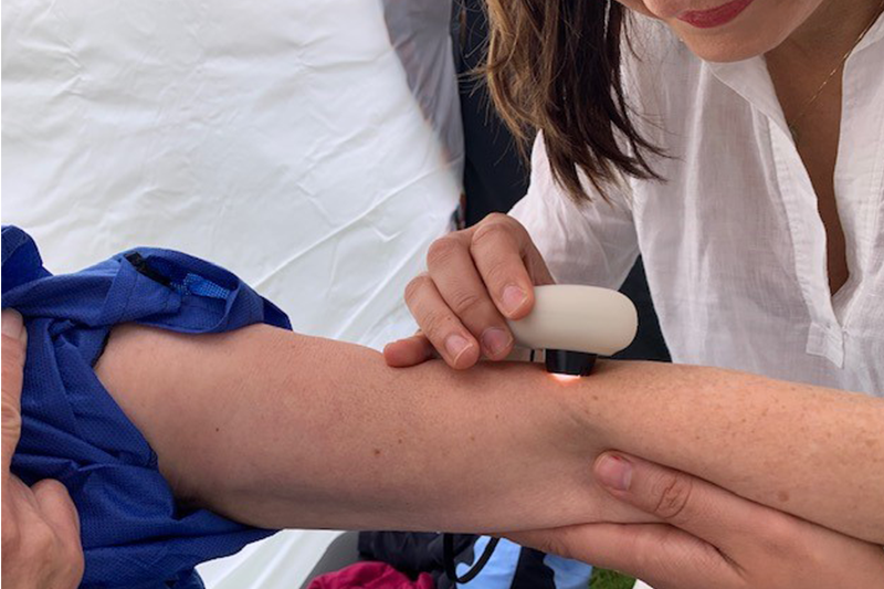 A woman holds back the sleeve of her dark blue silk shirt, exposing her arm to a nurse, who holds a pebble-shaped device over a freckle on the woman's arm. 