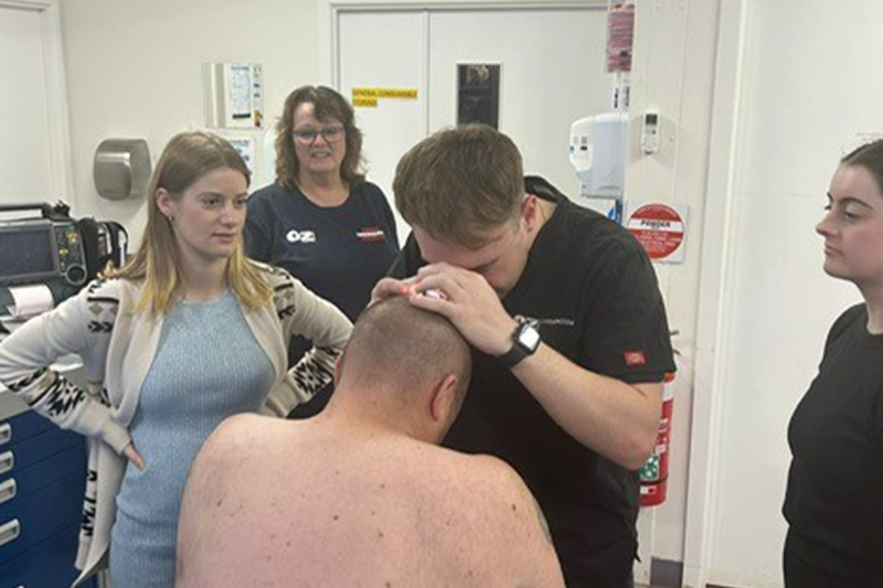 A group of four nurses gather in a small white treatment room. Three are wearing black t-shirts while the other, stationed to the left, is wearing a blue knit dress and white cardigan. Amongst the nurses sits a man. He has a closely shaved head and is shirtless. A nurse holds a pebble-shaped device up to the man's head, checking for moles and freckles that may be suspicious lesions 