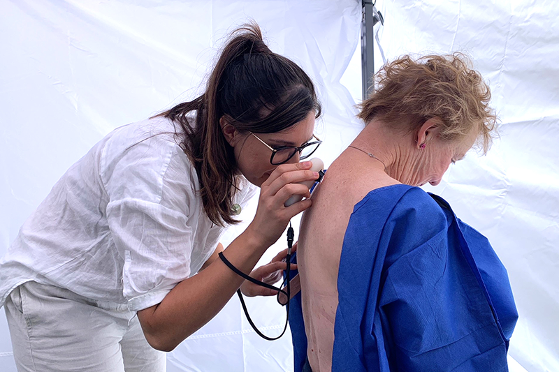 A medical professional, with dark brown hair, glasses, and white linen enesemble, is using a small dermoscope to examine a mole of an elderly lady's back. The lady's back is exposed but the remainder of her is covered in blue scrubs. They are both situated insight a white tent.