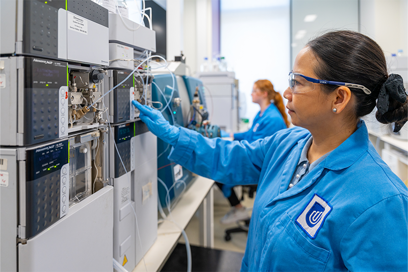 A woman with dark hair, wearing clear safety goggles and a blue lab coat, presses buttons on a tall metal machine within a lab