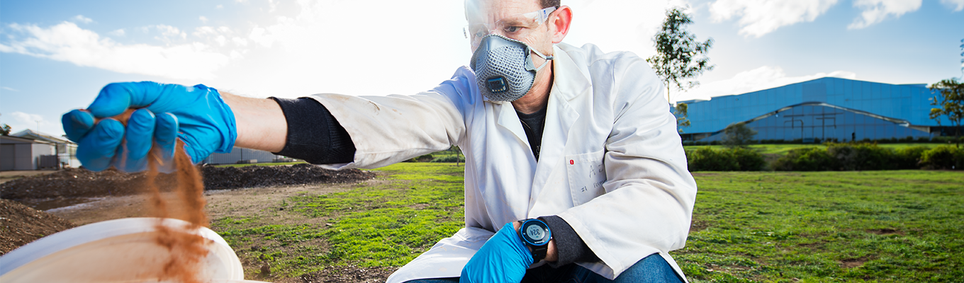 A researcher, wearing a black long sleeved t-shirt covered mostly by a white lab coat kneels in a green field. He is wearing a white face mask over his nose and mouth. He is also wearing bright blue plastic gloves. In his right hand, he is holding a fistful of red-brown dirt, some of which is falling back into a white bucket.