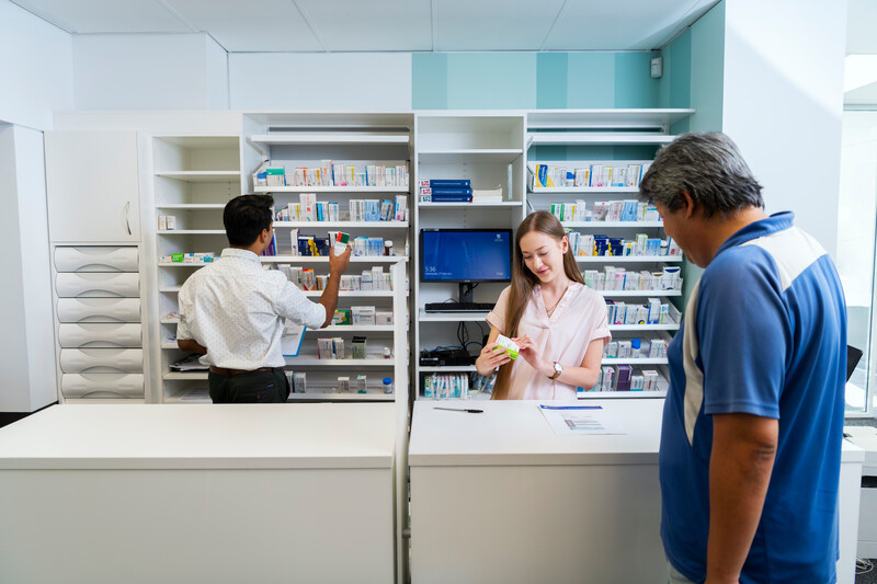 A man being served at a counter by a female pharmacy student. behind her are shelves of medications. Another male student is looking at the contents of the shelves