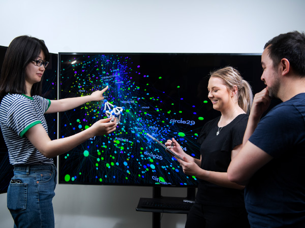 Three people gather around a TV screen. The screen has an image with a blackbakcground, and a spiderweb of interconnected dots and lines. The dots and lines are blue and green. 