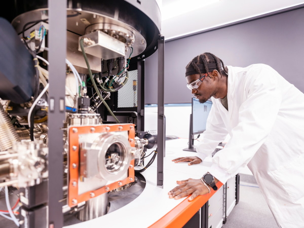 A student wearing a white lamb coat stands right of screen, leaning against a metal bench. He inspects a large metal machine that has a prominent circular shape.