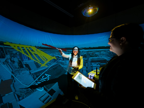 Two people stand in a dark room. The person to the right of screen is mostly in shadow, holding a clipboard. The other person stands in the middle of the screen, holding a pencil up to a projected image. The image, projected onto a curved wall, is taller than the two people, and shows a map. 