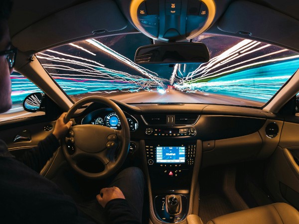 An interior of a car. The photo is taken from the middle passenger seat, peering over the shoulder of the driver. Neon strips of light can be seen through the car's windows and windshield, giving the impression of the car moving quite rapidly.