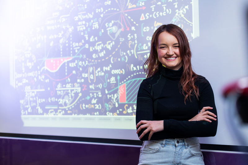 student smiling and arms crossed in front of a digital screen with engineering graphics