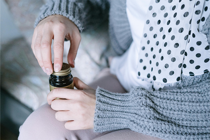 A hand, that appears to belong to a young woman, holds a small glass vial with a metal twist lid. She is wearing a wan blue cardigan and is sitting on a bed. 