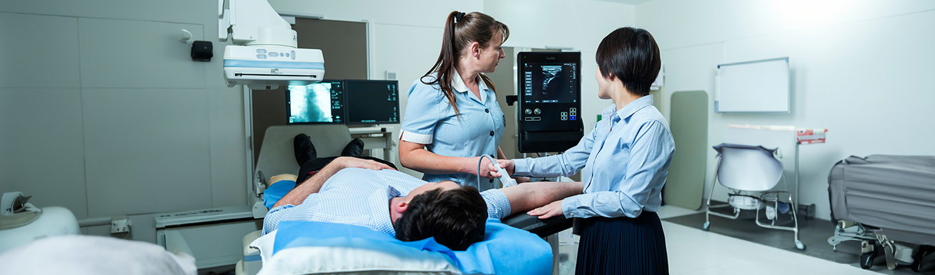 Two nurse practioners, dressed in light blue uniforms, perfrom an ultrasound on a patient's arm. Their veins are visible on the ultrasound screen. The patient lies in a bed, dressed in a sky blue gown. 