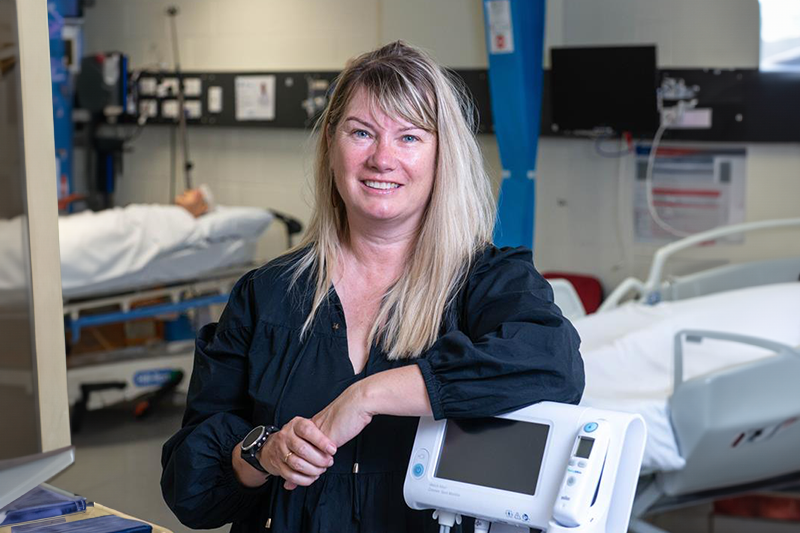 Professor Marion Eckert stands in a hospital near a heart monitoring machine. Behind her is a dummy patient in a bed. She has blonde hair and wears a dark coloured shirt. 