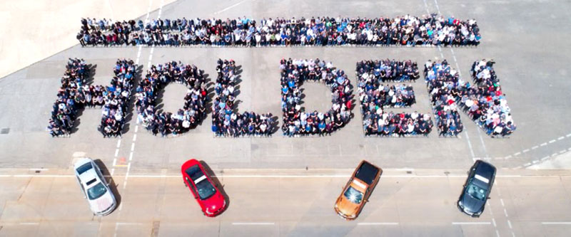Lots of people stand in groups to spell out the word HOLDEN with four cars in the foreground.