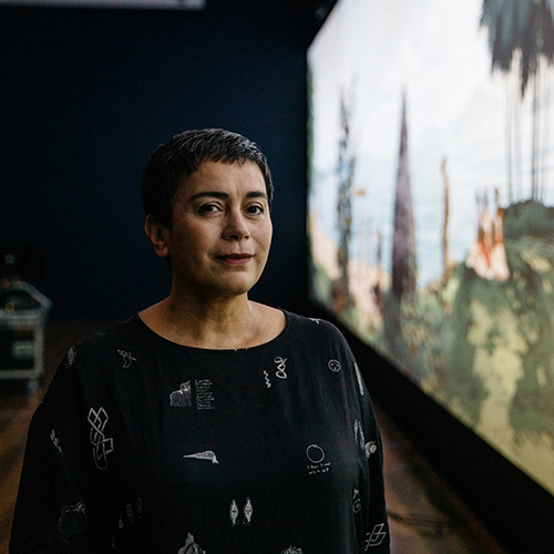 Portrait of Lisa Reihana standing next to her work, a New Zealand woman with short dark hair wearing a black top with white designs.jpg