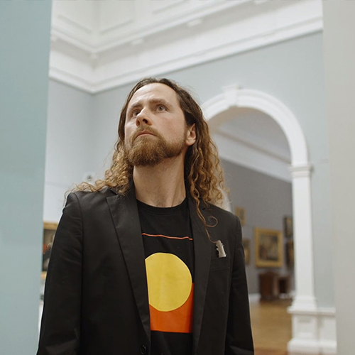 Portrait of Brad Darkson, an artist of Anglo Australian and Narungga background, standing in the Art Gallery of South Australia. Darkson has long, curly, light brown hair and wears a black t-shirt featuring the Aboriginal flag and  black jacket.jpg