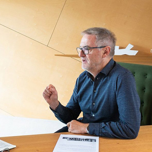 Profile view of John Wardle sitting at a desk speaking to someone outside of the frame. Wardle is a Caucasian man with short, grey hair wearing dark framed glases and a dark blue shirt.jpg