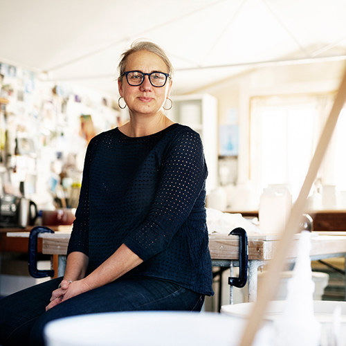 Portrait of Kirsten Coelho sitting on a table in her studio. Kirsten is a Caucasian female with blonde hair, tied back, wearing dark framed glasses, a dark top and indigo jeans.jpg