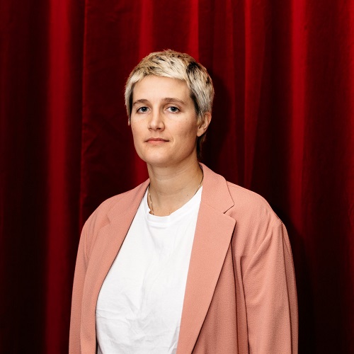 Portrait of Madison Bycroft standing in front of a red velvet curtain. Madison is a Caucasian person with short blonde hair wearing an peach coloured jacket over a white t-shirt.jpg