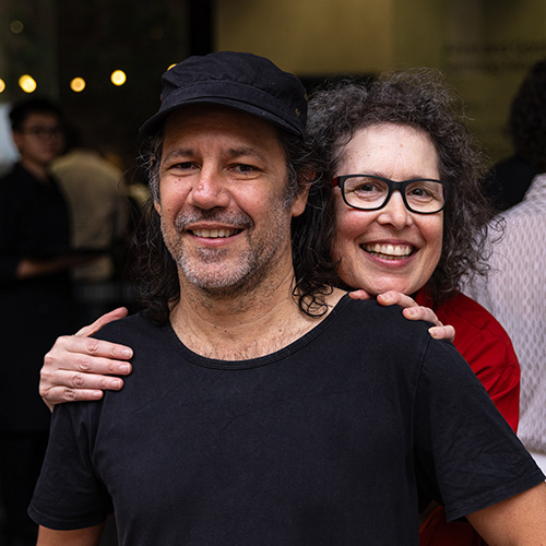 Photo of Molly Reynolds peeking over the shoulder of Archie Moore. Archie is an Aboriginal man with shoulder length, black hair wearing a black cap and t-shirt. Molly is a Caucasian woman with curly black hair wearing black framed glassess and a red shirt.jpg