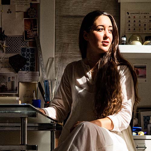 Portrait of Dana Awartani sitting on a chair in her studio. Dana is an Arabic woman with long, dark hair wearing a long white dress.jpg