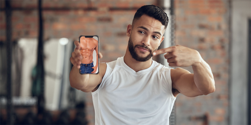 Young Man in a gym holding up an image of the perfect six-pack on his phone