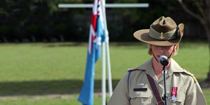 A young female Australian Army officer wearing full ceremonial uniform, traditional Sam Browne belt and the Australian Defence Medal gives an emotional speech to the community at the Coomera ANZAC Day remembrance ceremony. The lady (one person) is the focal point of the image, but a memorial cross and the Royal Australian Air Force (RAAF) flag can be seen flying at half-mast in the background.